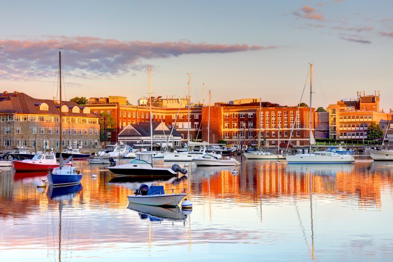 Boats and yachts are docked in a calm harbor, reflecting in the water, with red brick buildings and a partly cloudy sky illuminated by warm sunlight in the background.