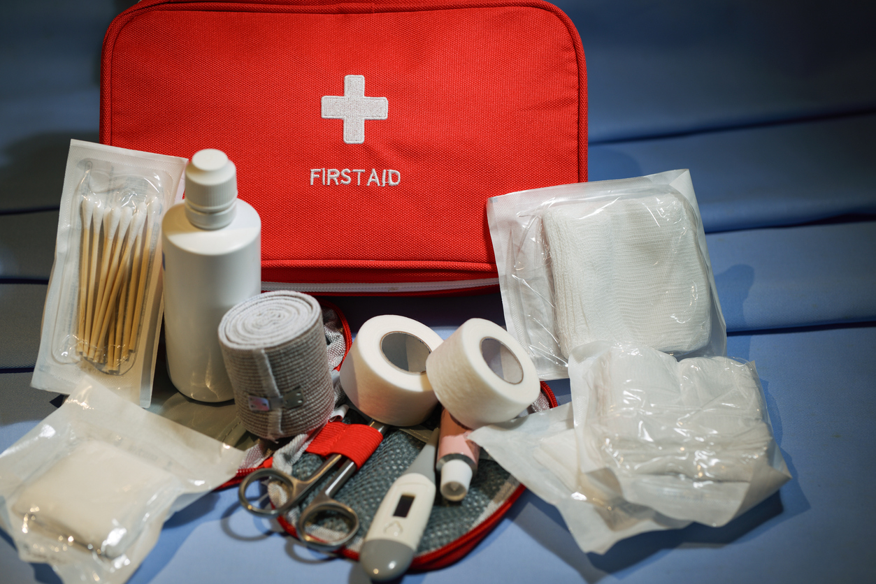 A red first aid kit with a white cross symbol is open, displaying medical supplies including bandages, gauze pads, adhesive tape, scissors, cotton swabs, a bottle, tweezers, and a digital thermometer.