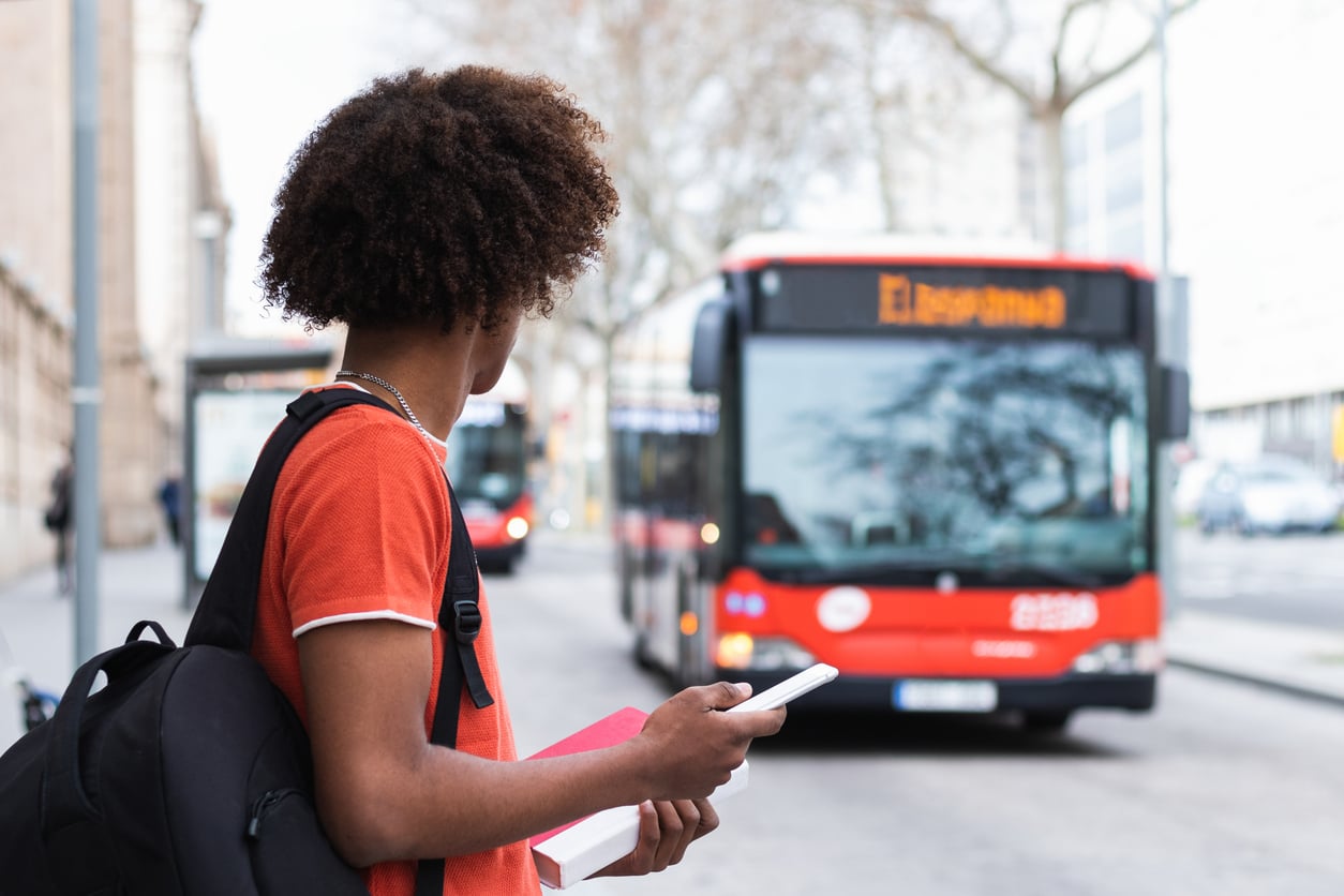 A person with curly hair, wearing an orange shirt and carrying a backpack, stands at a bus stop holding a phone and notebook, looking at an approaching red and white city bus.