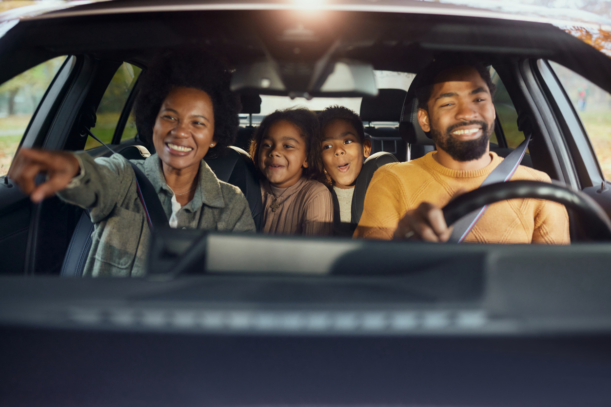 A smiling family of four sits in a car. The father is driving, the mother is pointing excitedly, and their two children are sitting happily in the back seat, looking ahead.
