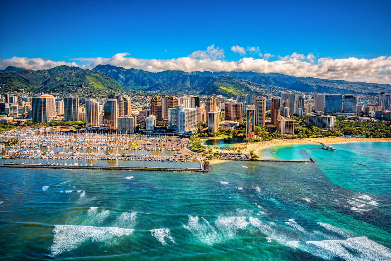 Aerial view of Waikiki Beach in Honolulu, Hawaii, with turquoise ocean waves, high-rise hotels, palm trees, and lush green mountains in the background under a bright blue sky.