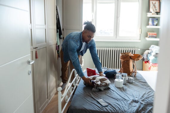 A person is packing clothes and essentials onto a bed in a bright bedroom, preparing a brown backpack. Various items, including a water bottle and rolls of toilet paper, are spread out on the bed.
