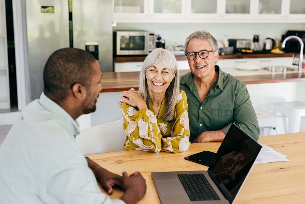 An older couple smiles while sitting at a kitchen table with a younger man. A laptop, smartphone, and papers are on the table, and the kitchen is visible in the background.