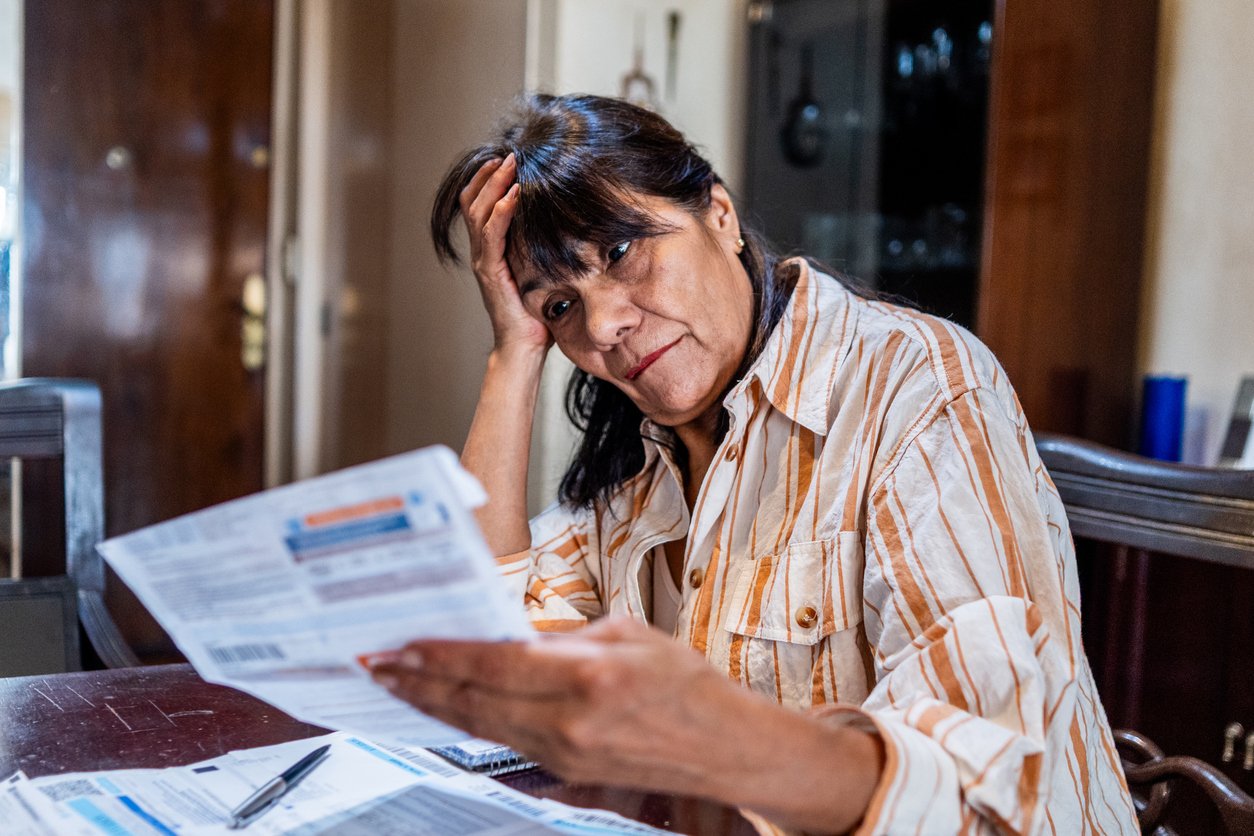A woman sits at a table holding and looking worriedly at bills, with her other hand resting on her head. Papers and envelopes are spread out in front of her, suggesting concern over finances.