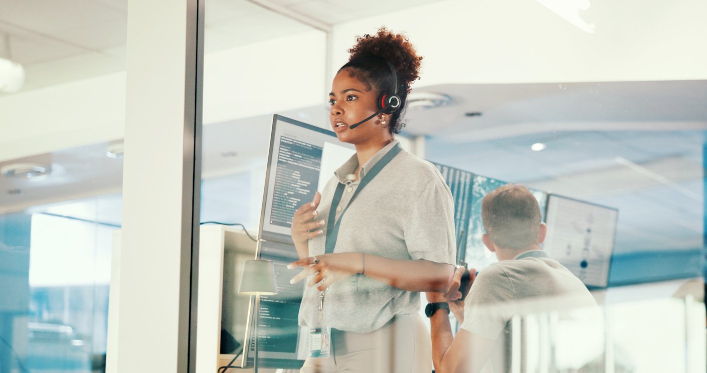 A woman wearing a headset stands in front of computer monitors, speaking and gesturing in a modern office with glass walls. Another person is seated nearby, partially visible.
