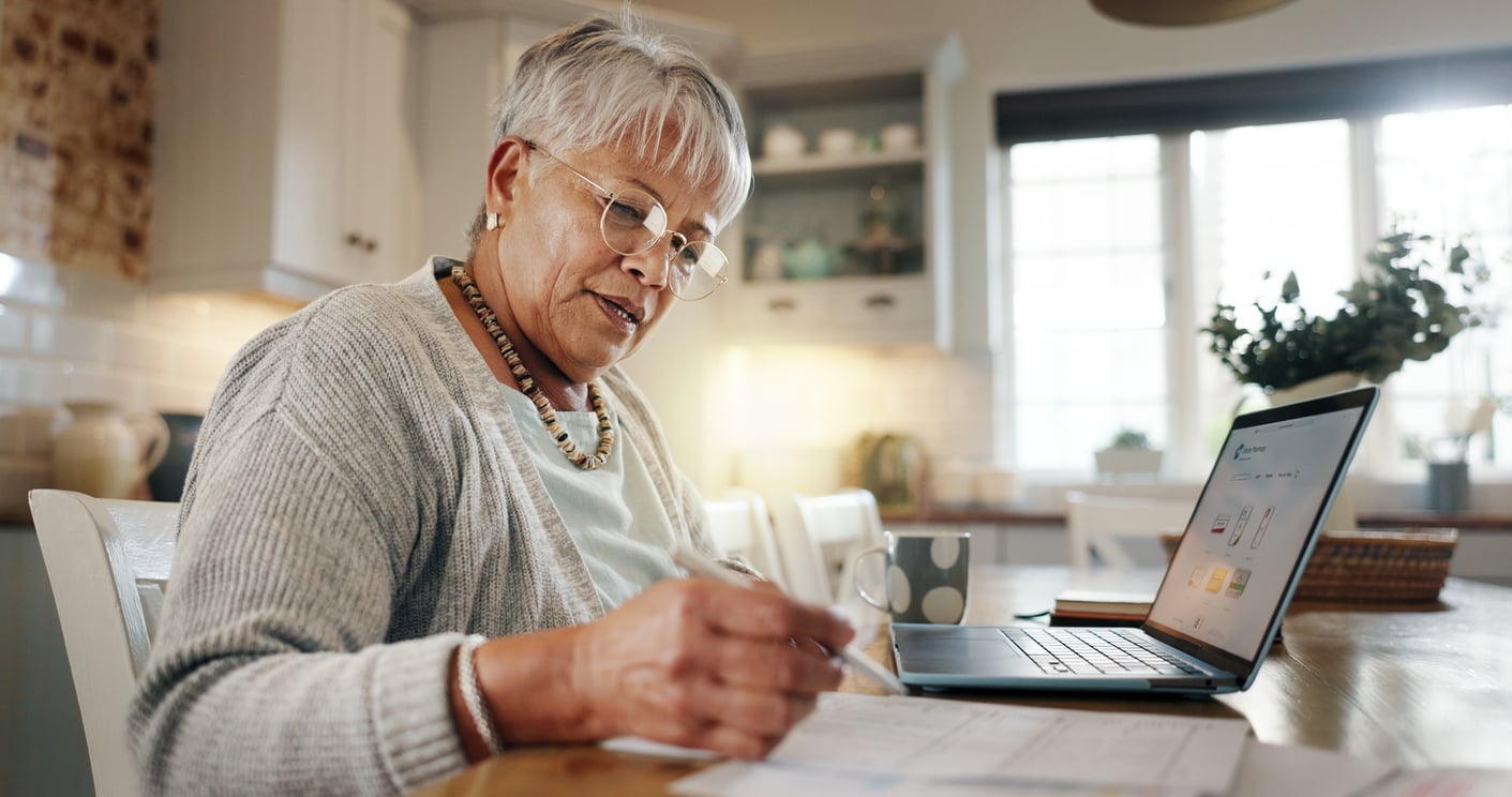 An older woman with short gray hair and glasses sits at a kitchen table, writing on papers with a pen next to an open laptop. The kitchen is bright with natural light.