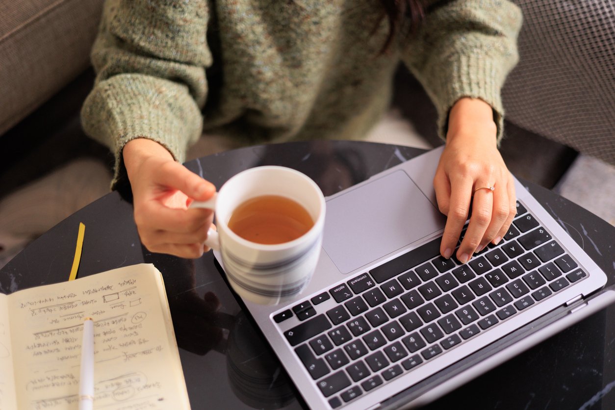 A person wearing a green sweater sits at a table, holding a mug of tea in one hand and using a laptop with the other. A notebook with handwritten notes and a pen are on the table beside the laptop.