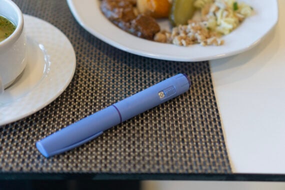 A blue pen rests on a woven gray placemat next to a white cup and a plate of food with rice, vegetables, and meat.