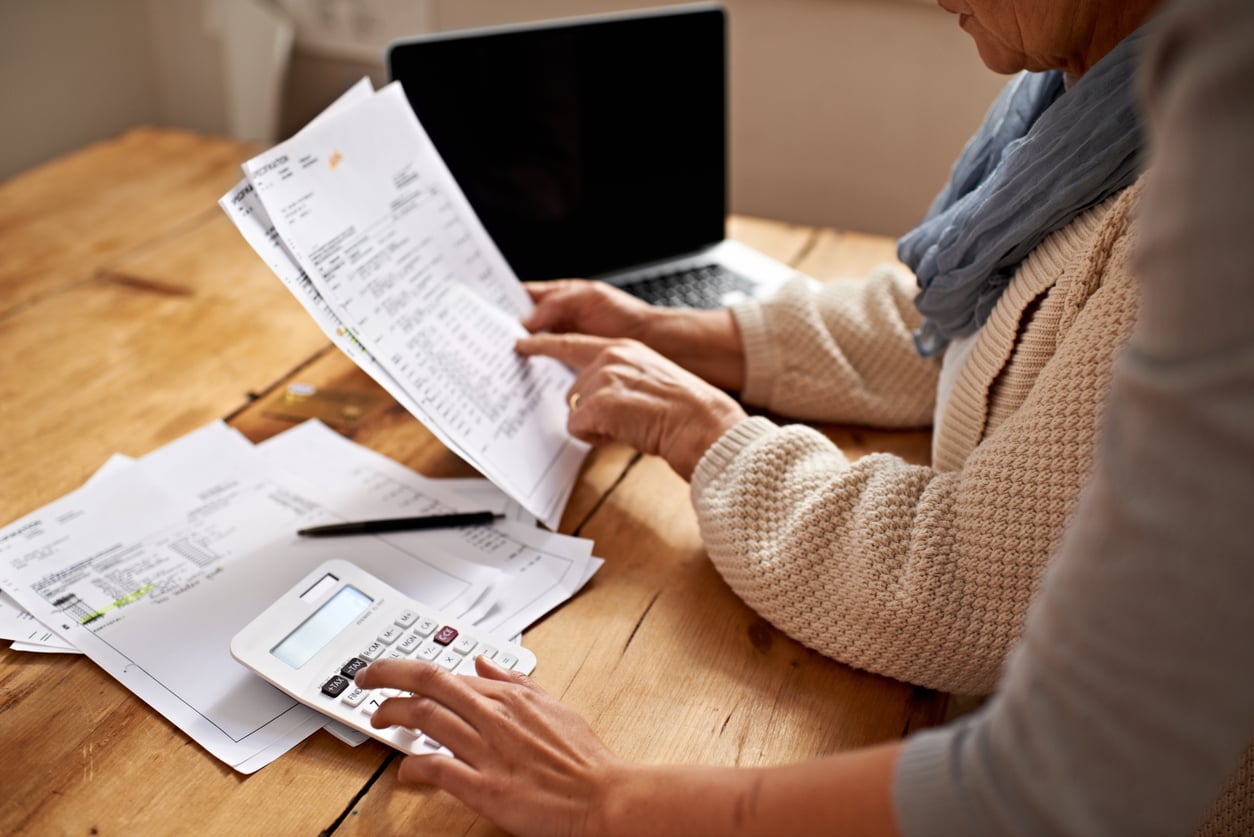 Two people sit at a wooden table reviewing financial documents and using a calculator, with a laptop open in the background. One person points to a page while the other enters numbers on the calculator.