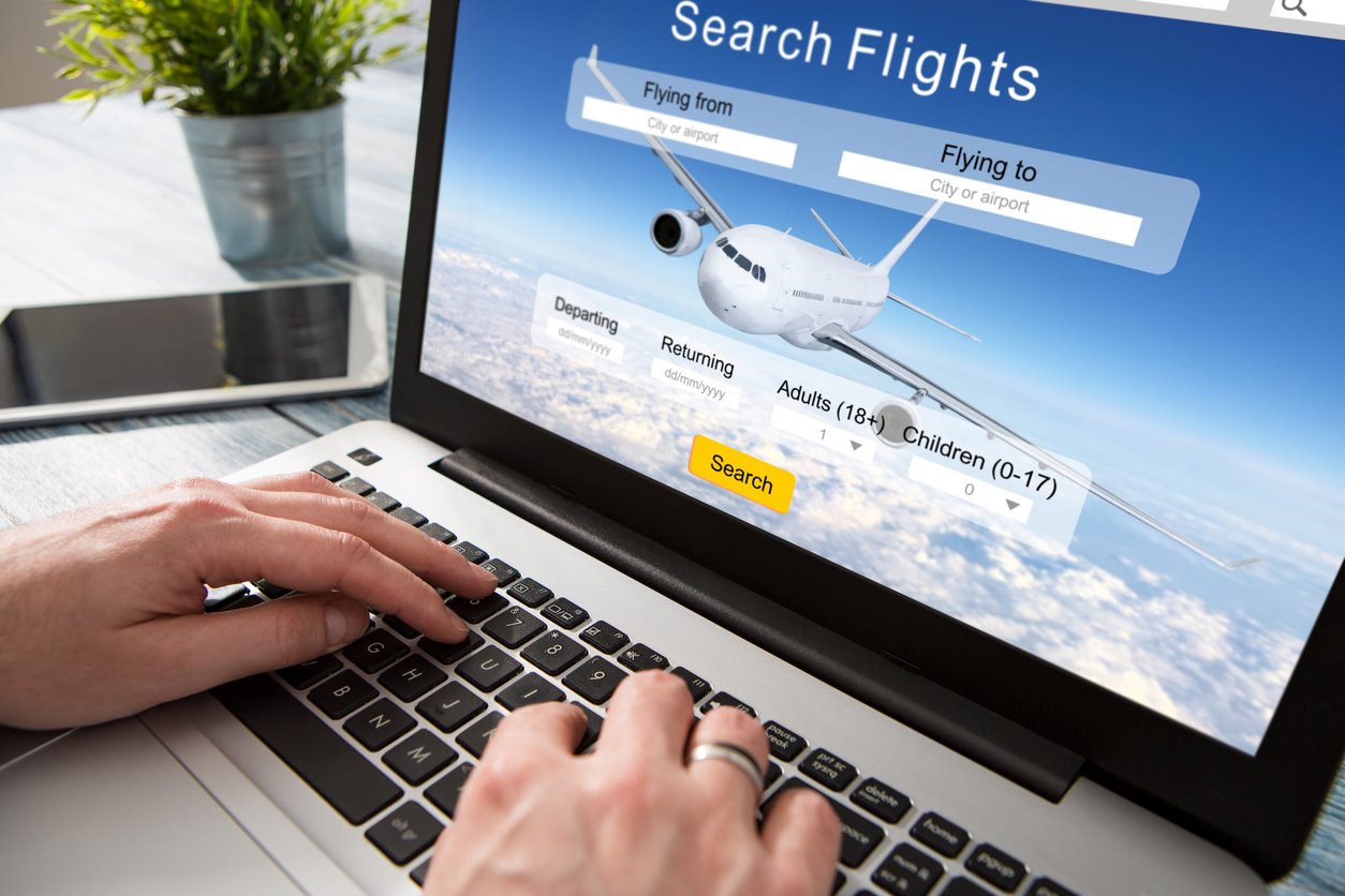 Hands typing on a laptop keyboard with a flight search website open on the screen, showing options to enter departure and destination cities, travel dates, and number of passengers.