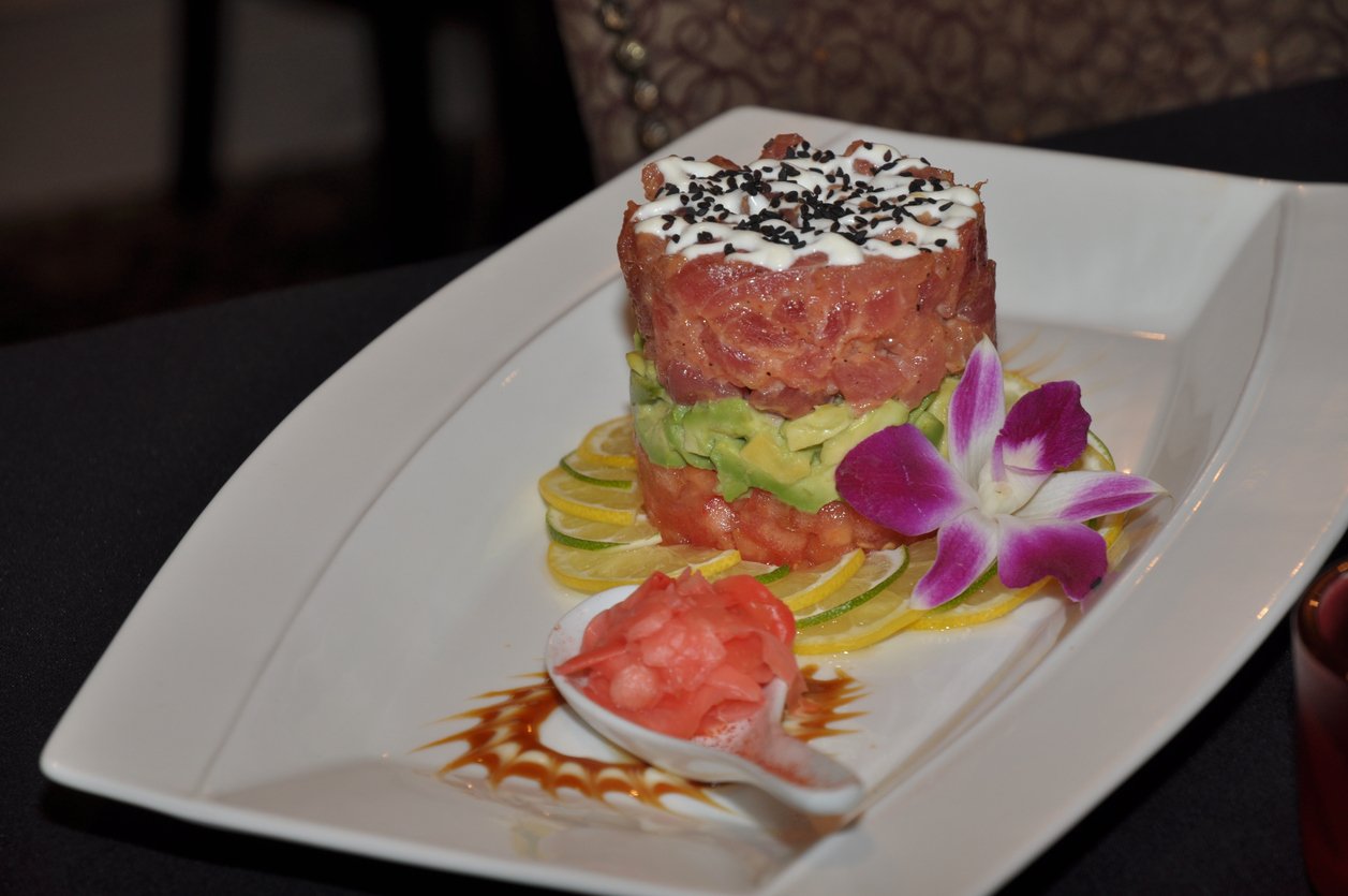 A layered tower of tuna tartare, avocado, and a creamy topping garnished with black and white sesame seeds, served on lemon slices with pickled ginger and a purple orchid on a white plate.