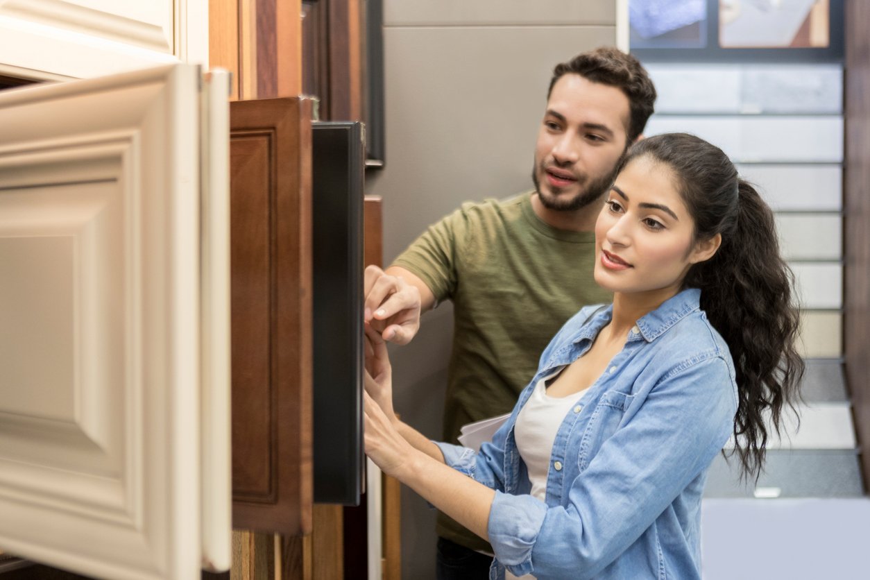 A man and woman look at and discuss cabinet door samples in a showroom. The woman touches one of the sample doors while the man points at another display. They appear to be considering options for a renovation.