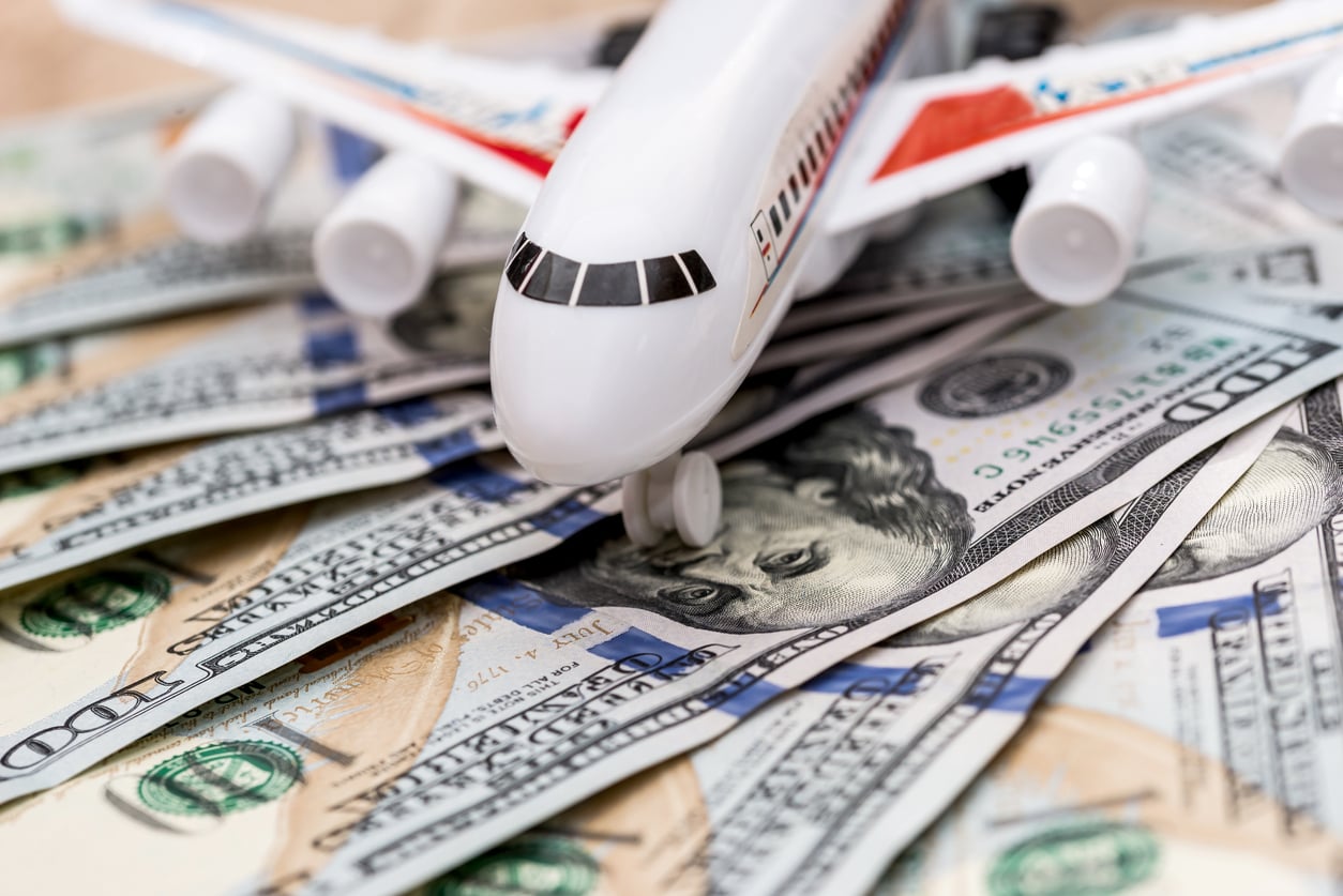 A model airplane sits atop scattered US hundred-dollar bills, symbolizing the concept of travel expenses or the cost of air travel.