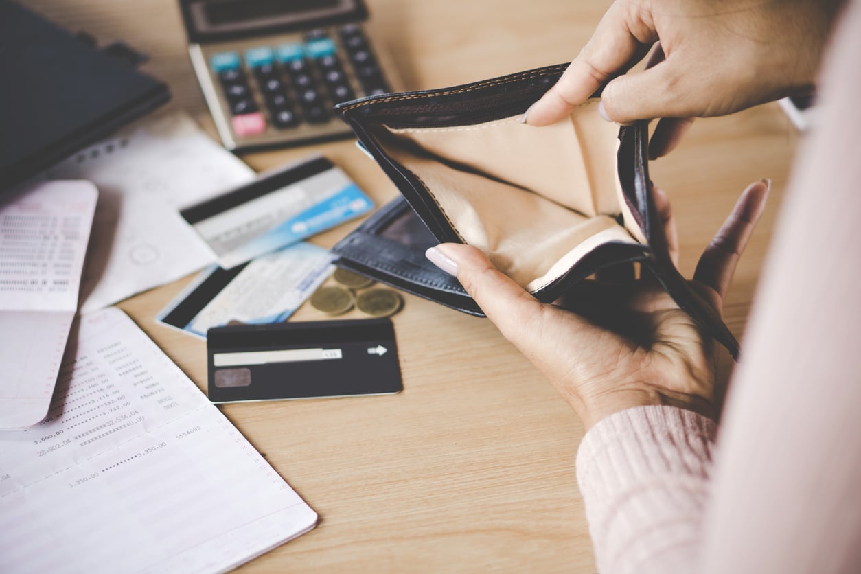 A person holds open an empty wallet over a desk with credit cards, coins, documents, a calculator, and a bank statement, suggesting financial difficulties.