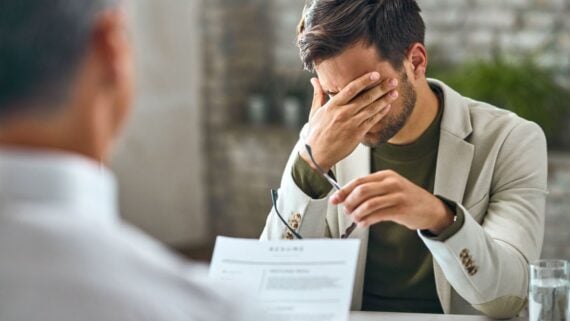 A man in a beige blazer sits at a desk, covering his face with one hand and holding eyeglasses in the other, appearing stressed during a meeting with another person who holds a document.