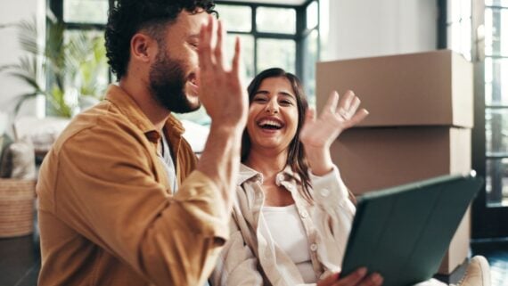 A smiling man and woman sit together indoors, waving at a tablet screen. Cardboard boxes are stacked behind them, suggesting they are in the process of moving. Bright light fills the room.