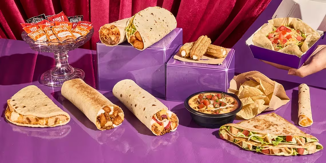 A variety of Taco Bell menu items, including wraps, tacos, nachos with cheese, churros, and sauce packets, are displayed on a shiny purple surface with a red curtain in the background.
