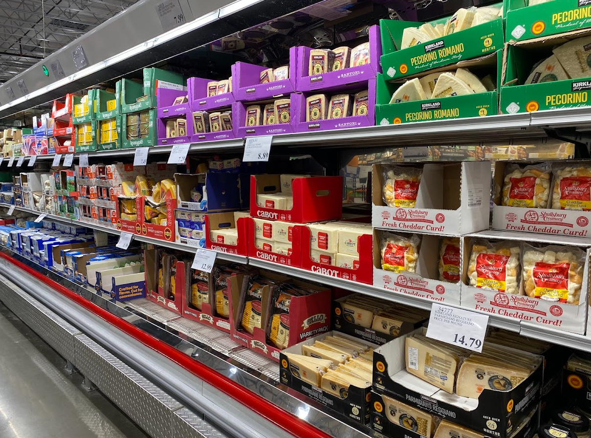 Shelves in a grocery store display a variety of packaged cheeses, including cheddar, pecorino romano, and mozzarella, in different brands and sizes. Each cheese is stacked neatly in boxes or wrapped in plastic.