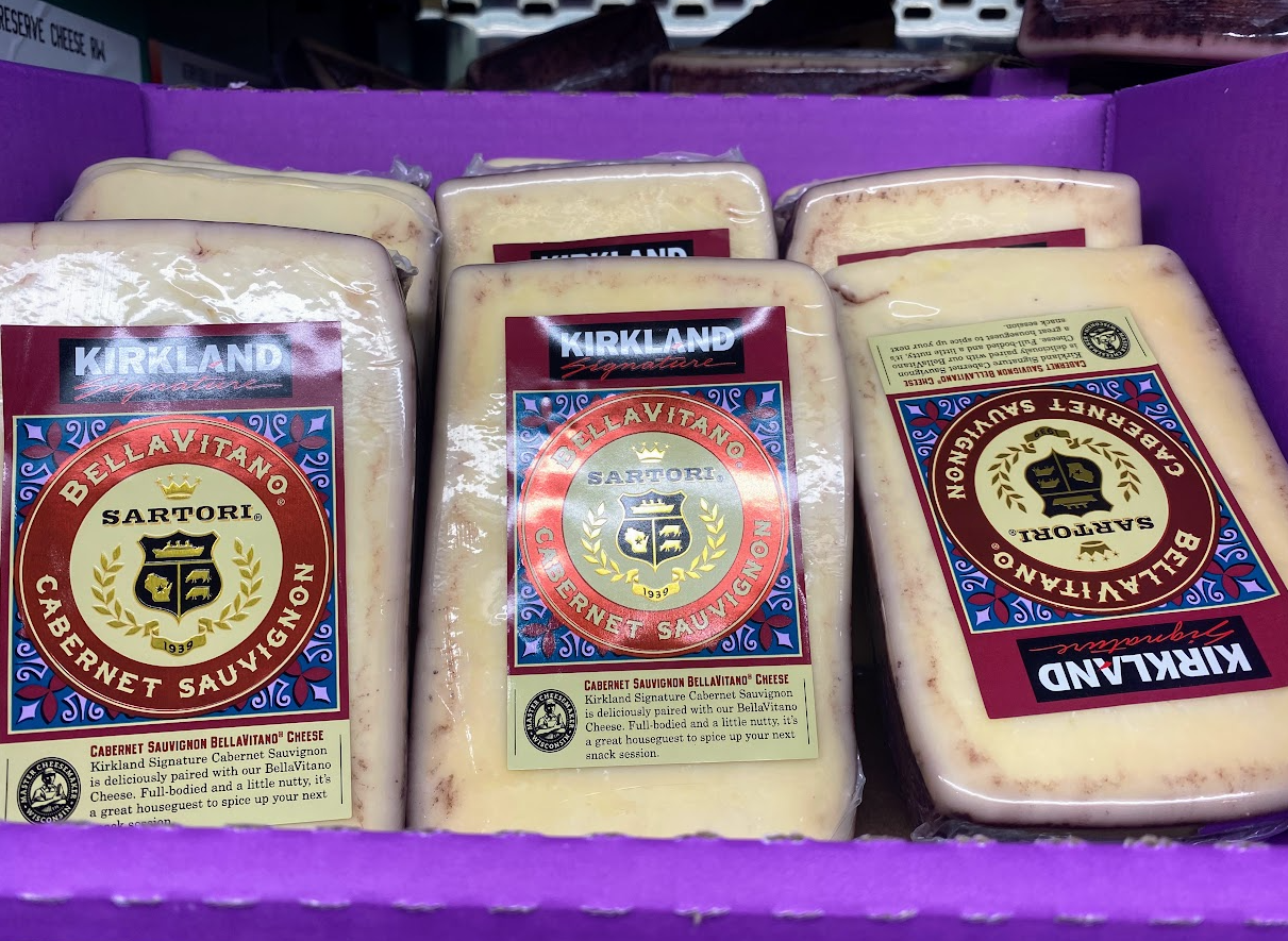 A display of Kirkland Bellavitano Cabernet Sauvignon cheese wedges in clear packaging, arranged in a purple box. Each cheese block features a decorative label with gold, red, and blue accents.