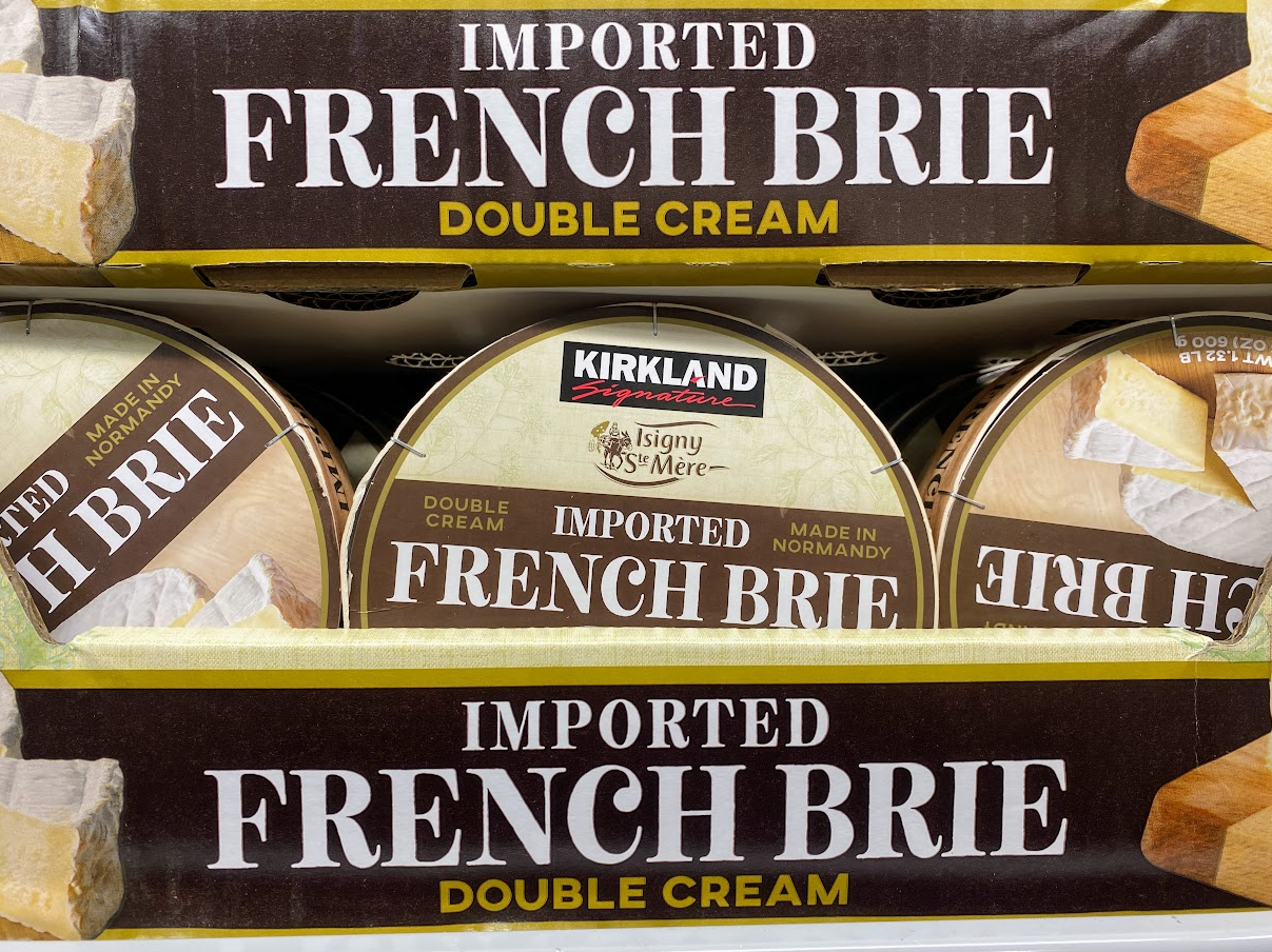 Close-up of stacked packages of Kirkland Signature imported French Brie double cream cheese, labeled “Made in Normandy.” The packaging is brown and tan with white and yellow text.