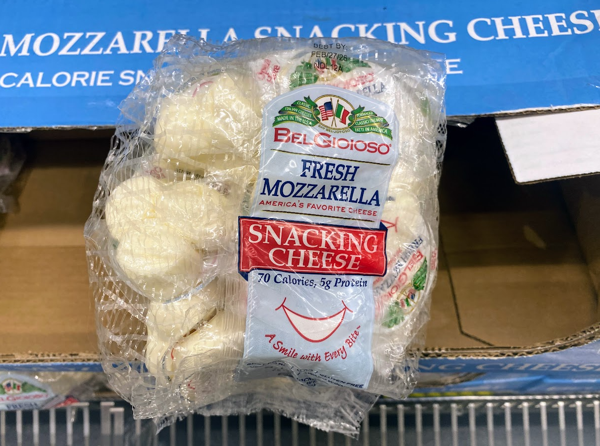 A package of BelGioioso Fresh Mozzarella Snacking Cheese sits in front of a blue box. The clear packaging shows small mozzarella balls and lists nutritional info: 70 calories, 5g protein.