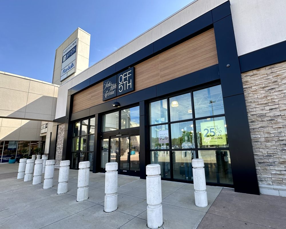 Entrance to a Saks OFF 5TH store, featuring large glass doors, promotional posters offering 25% off, and a row of white bollards on a sunny day. The building has tan brick and wood paneling accents.