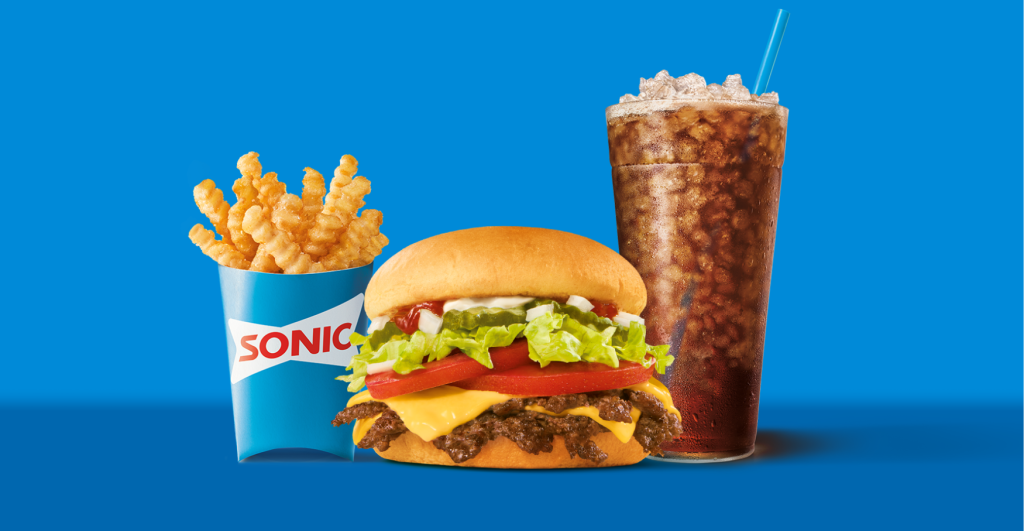 A new fast-food menu meal from Sonic features crinkle-cut fries in branded packaging, a cheeseburger with lettuce, tomato, and cheese, plus a large soda with ice in a clear cup and blue straw, all set on a blue background.