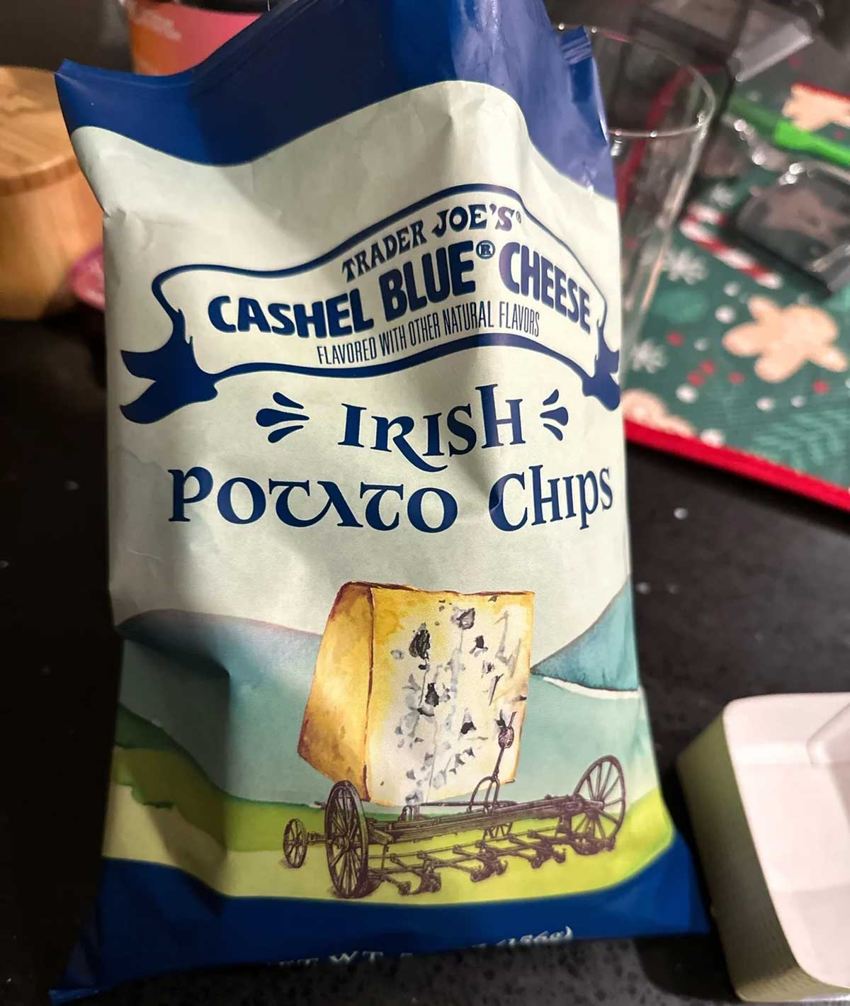A bag of Trader Joe’s Cashel Blue Cheese Irish Potato Chips sits on a counter. The packaging features an illustration of a cheese wheel on a wooden cart in a green field.