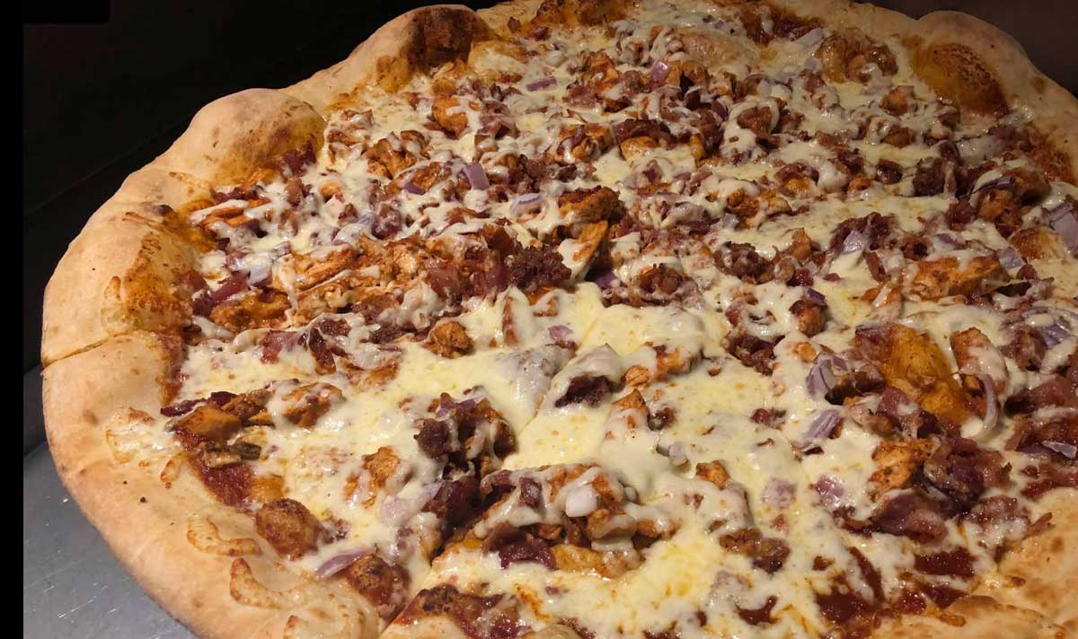 A close-up view of a large pizza topped with melted cheese, pieces of chicken, bacon bits, and red onions on a thick, golden-brown crust—just like the hearty east coast cheap pizza slices everyone loves.