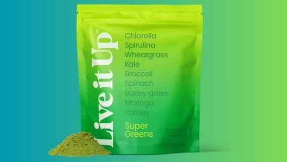 A green and yellow pouch labeled "Live it Up Super Greens" stands upright with a pile of green powder beside it. Ingredients like chlorella, spirulina, kale, and spinach are listed on the front of the pouch.