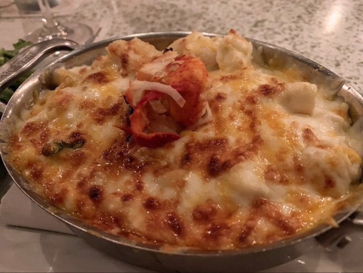 A metal dish filled with a baked, cheesy seafood casserole topped with melted, golden-brown cheese and a lobster claw garnish, set on a table.