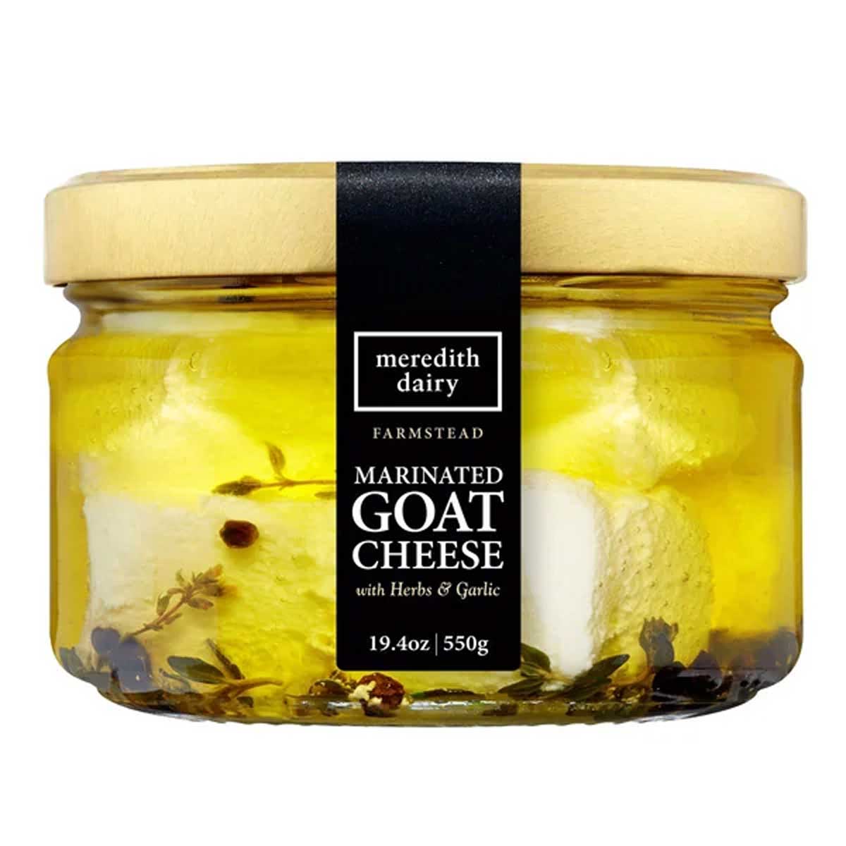 A glass jar of Meredith Dairy Farmstead Marinated Goat Cheese with herbs and garlic, filled with oil and visible herbs. The jar has a wooden lid and a black label displaying the product name and weight.