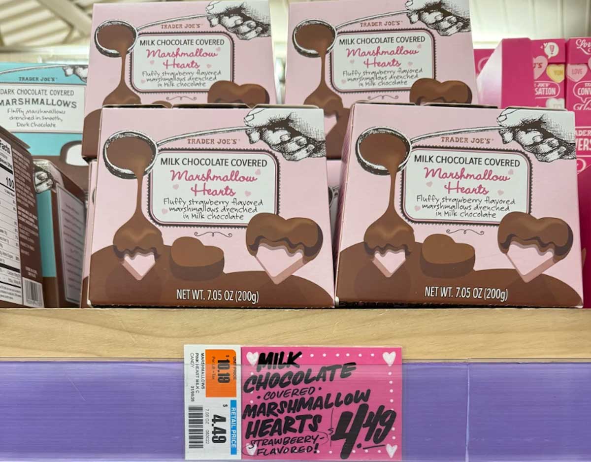 Trader Joe’s boxes of milk chocolate covered marshmallow hearts are stacked on a wooden shelf above a pink price tag labeled $4.49. The boxes feature images of chocolate heart shapes and dripping chocolate.