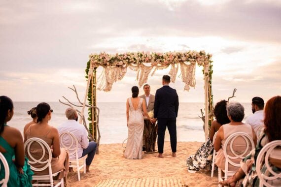 A couple stands facing an officiant under a floral arch on a sandy beach, surrounded by seated guests, with the ocean and a cloudy sky in the background.