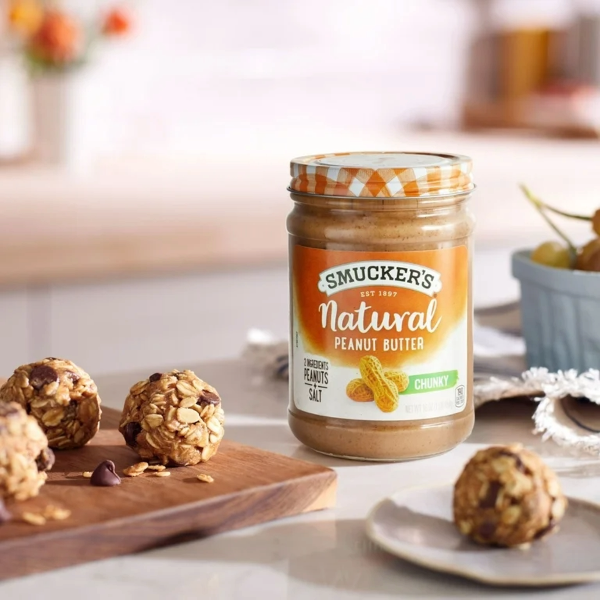 A jar of Smucker's Natural Chunky Peanut Butter sits on a kitchen counter next to a plate and a wooden board with homemade oatmeal energy balls. A bowl of fruit is visible in the background.