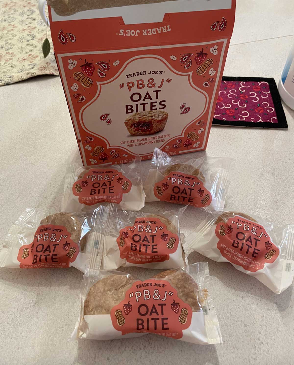 Six individually wrapped Trader Joe’s PB&J Oat Bites are arranged in front of an open box with the same branding on a countertop. The packaging is decorated with peanut and strawberry illustrations.
