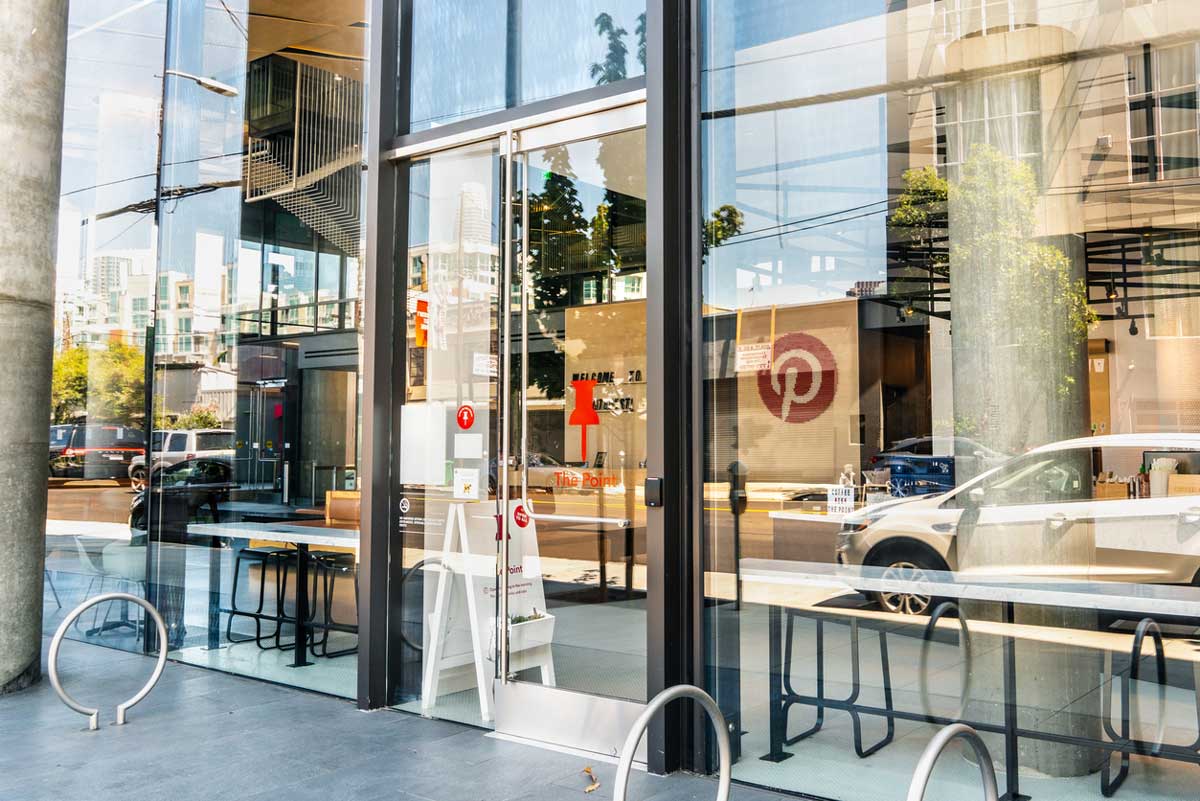 Glass storefront with a red arrow sign on the door, bike racks, long tables, and benches outside. Reflections of city buildings and cars—perhaps impacted by the 2026 layoffs—are visible in the large windows.