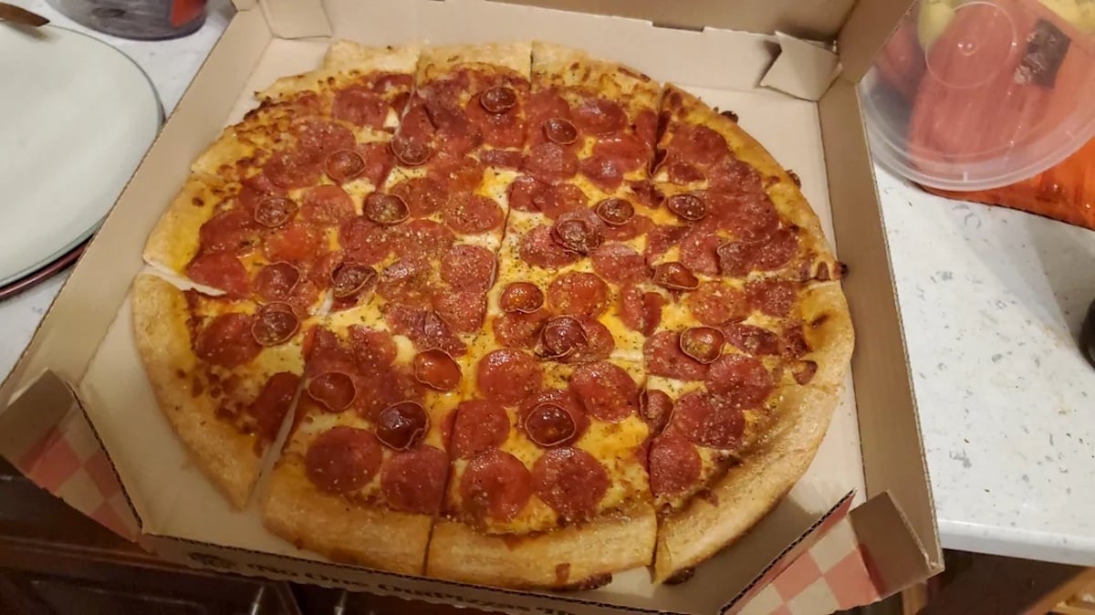 A pepperoni pizza cut into square slices sits in an open cardboard box on a kitchen counter. The pizza has a golden crust and is topped with pepperoni and sprinkled seasoning.