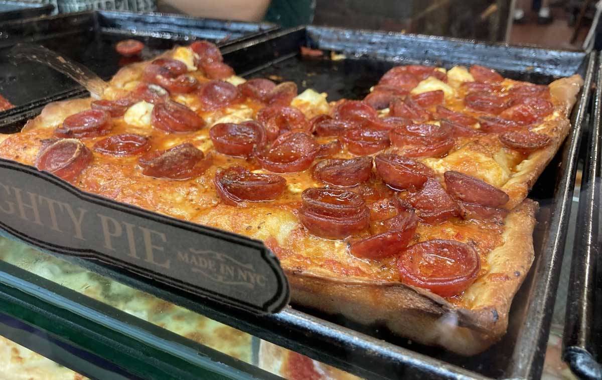 A close-up of a rectangular pepperoni pizza with crispy edges and melted cheese, displayed in a glass case at a pizzeria known for classic East Coast cheap pizza slices.