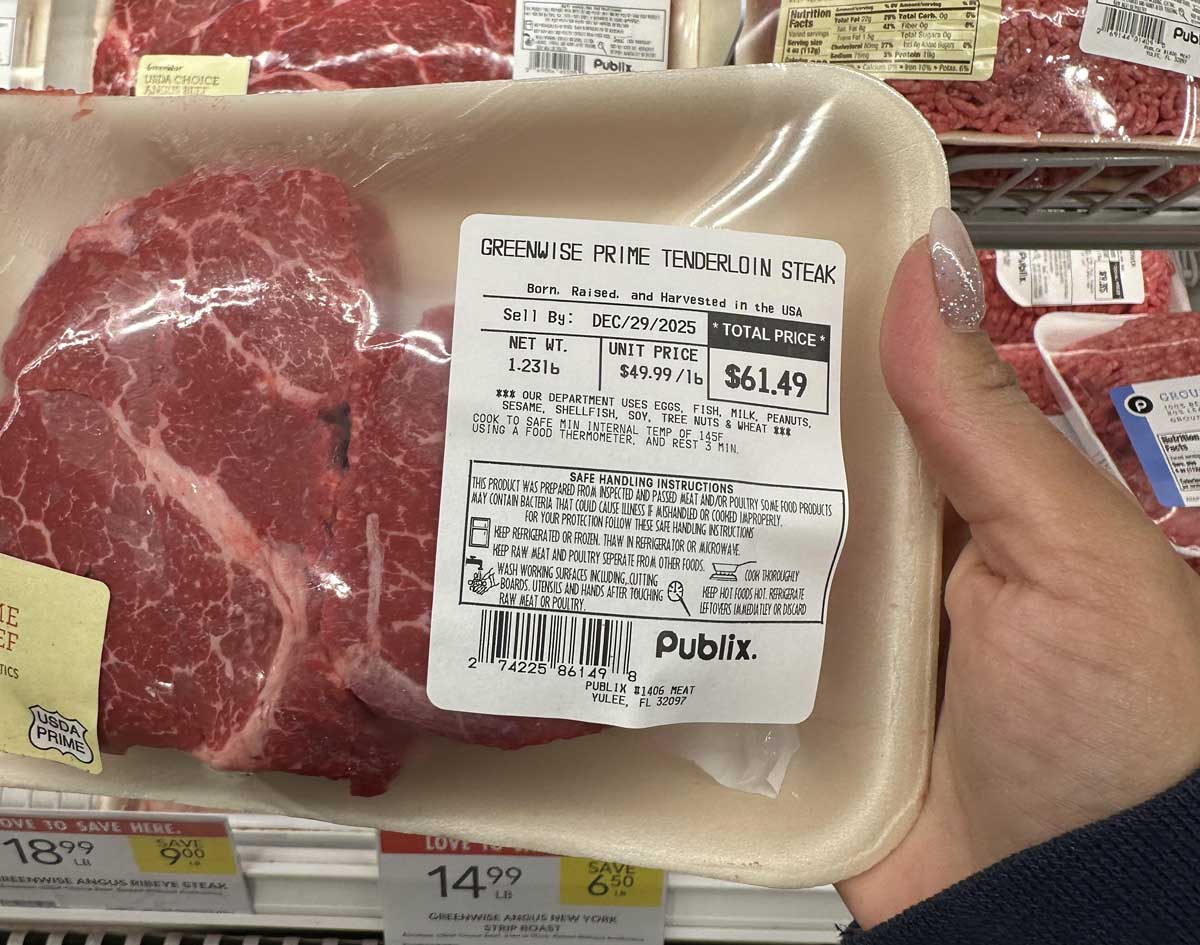 A hand holds a package of GreenWise Prime Tenderloin Steak from Publix, weighing 1.23 lbs and priced at $61.49. The label details weight, price per pound, and safe handling instructions. Other meat packages are visible in the background.