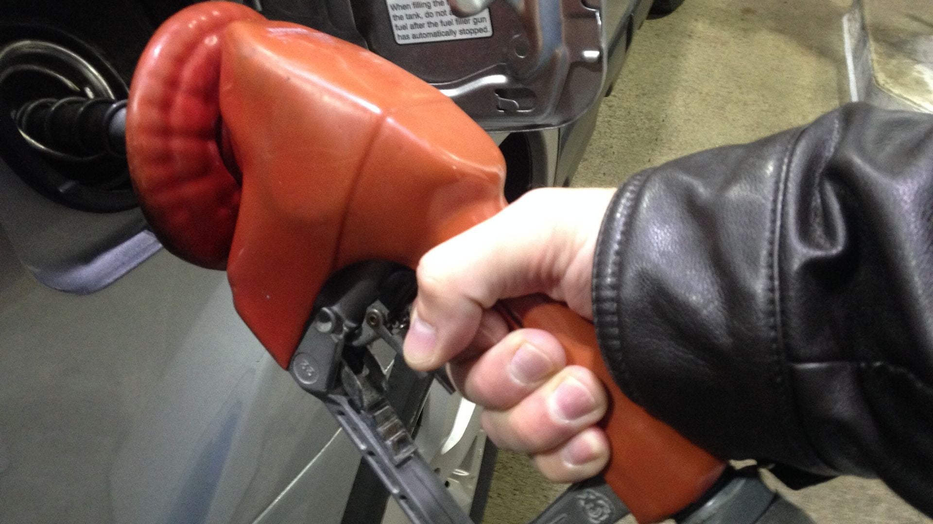 A person wearing a black leather jacket holds an orange gas pump nozzle while refueling a vehicle at a gas station.