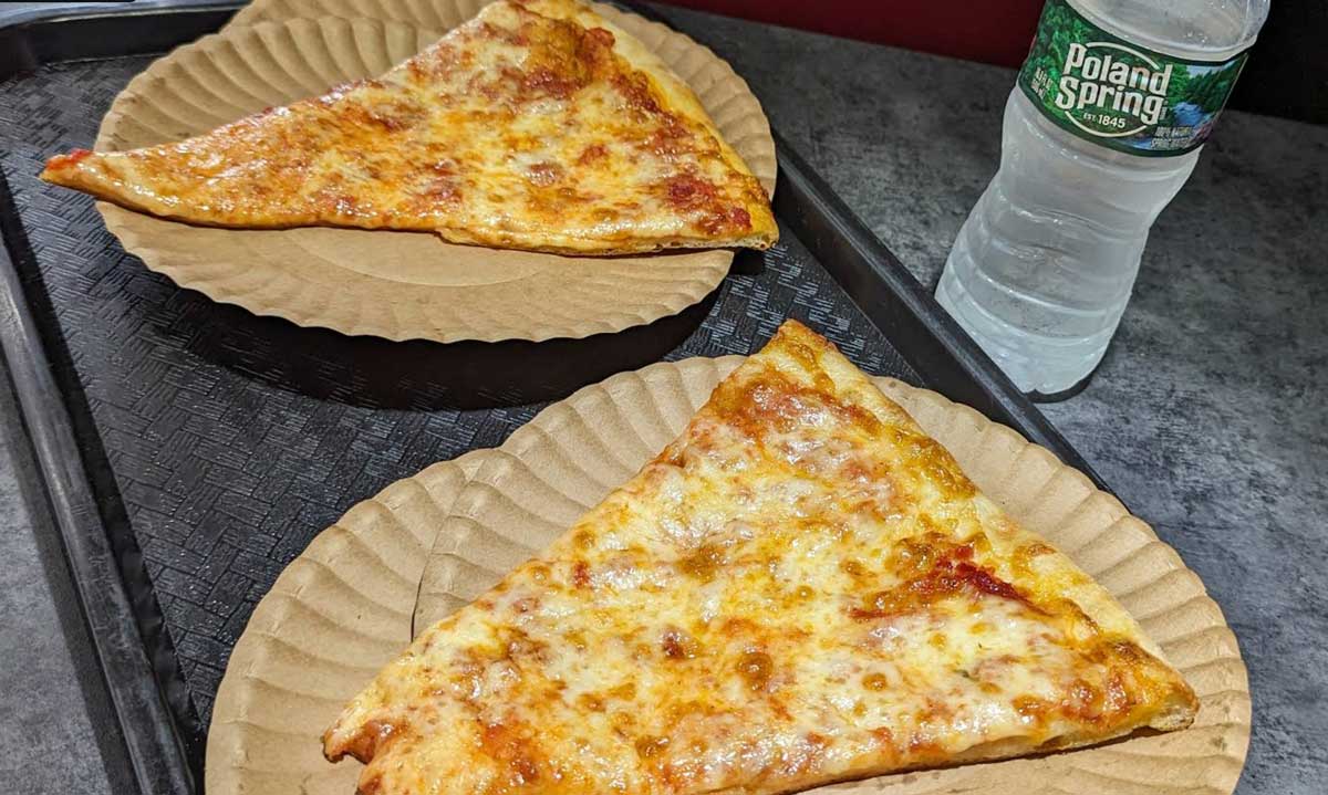Two slices of classic East Coast cheap pizza on brown paper plates, served on a black tray with a bottle of Poland Spring water to the upper right, all set atop a gray tabletop.
