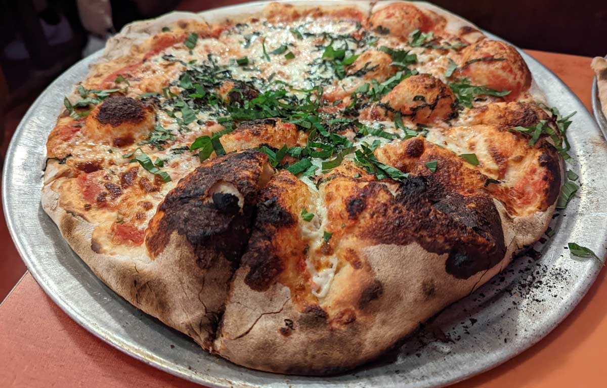 A close-up of a wood-fired pizza on a metal tray, reminiscent of classic East Coast cheap pizza slices, topped with melted cheese, tomato sauce, and fresh chopped herbs, with a bubbly, charred crust.