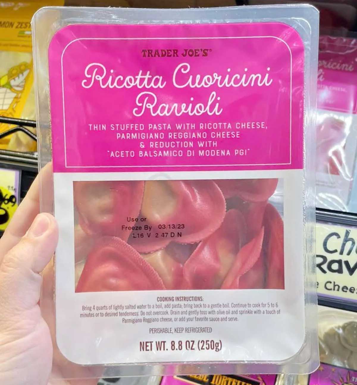 A hand holds a package of Trader Joe's Ricotta Cuoricini Ravioli, featuring heart-shaped pink pasta stuffed with ricotta and Parmigiano Reggiano cheese, displayed in a refrigerated store section.