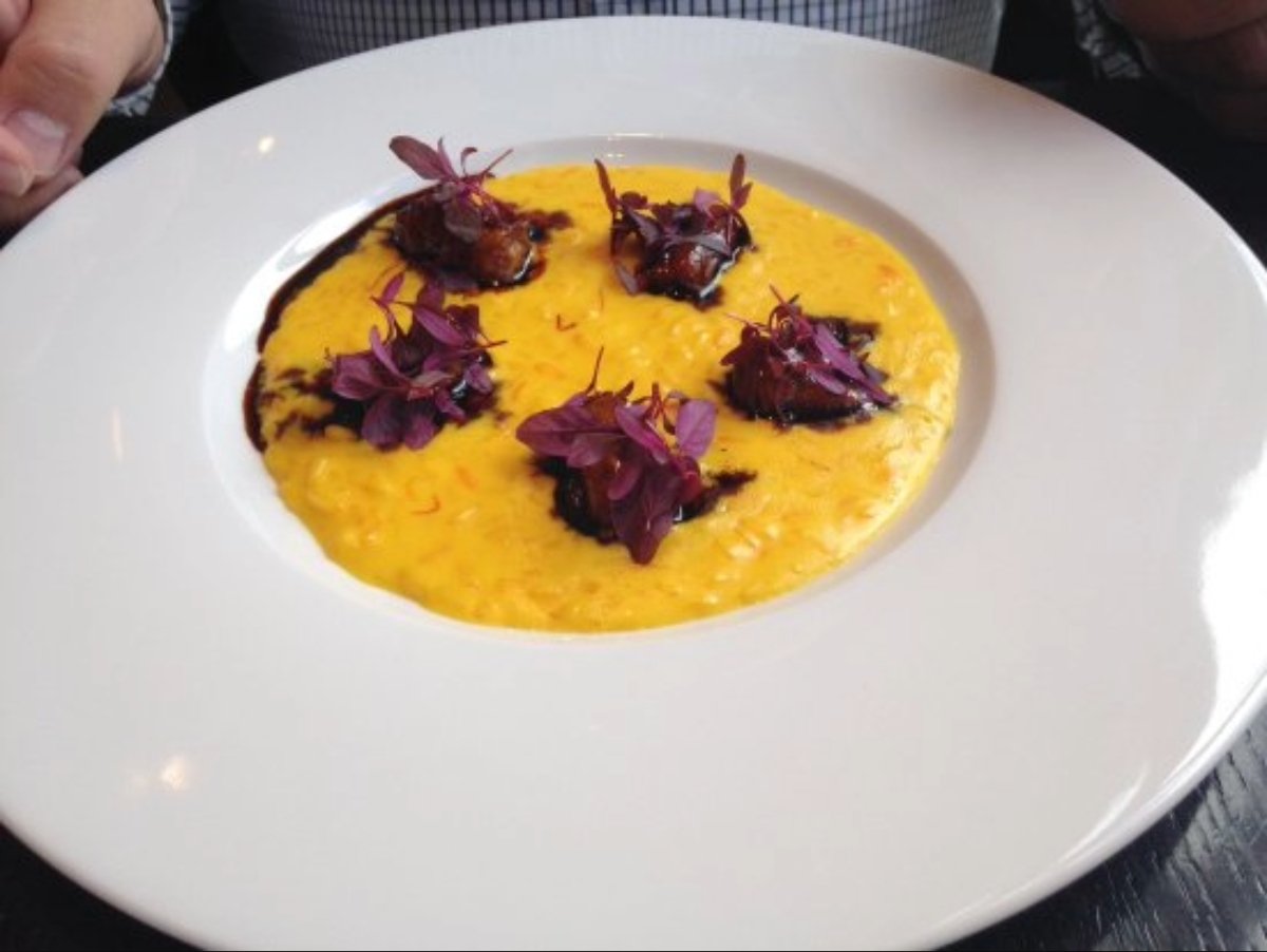 A white plate with creamy yellow risotto, topped with four small portions of a dark sauce and garnished with purple microgreens. A person’s hands are visible near the plate.
