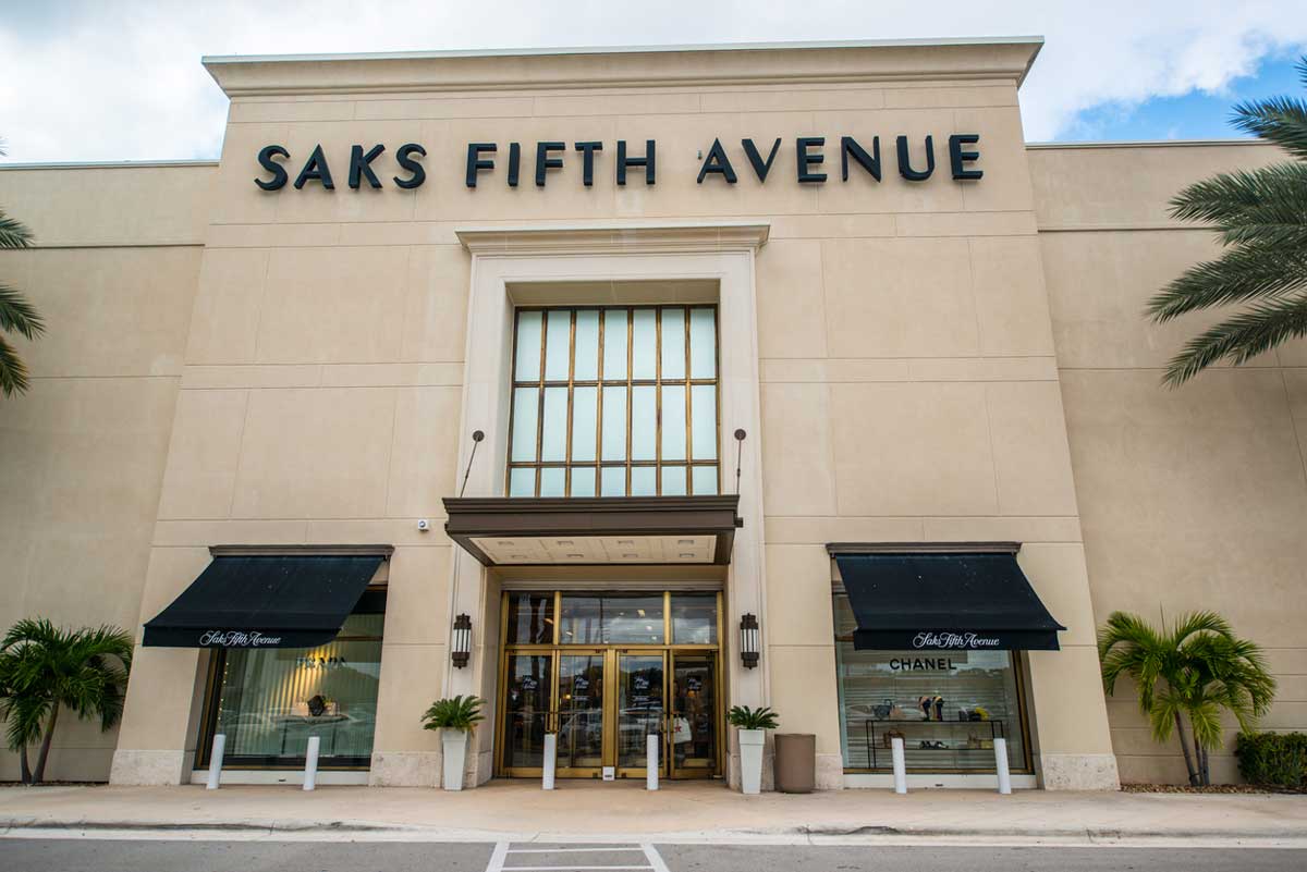 The exterior of a Saks Fifth Avenue store with large black signage, glass doors, two black awnings, display windows, palm trees, and a cloudy sky in the background amid news of 2026 layoffs.