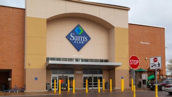 The entrance of a Sam&rsquo;s Club store with large blue signage above glass doors. Yellow posts line the front, and two red stop signs are visible to the right, near a pharmacy sign and a play area.