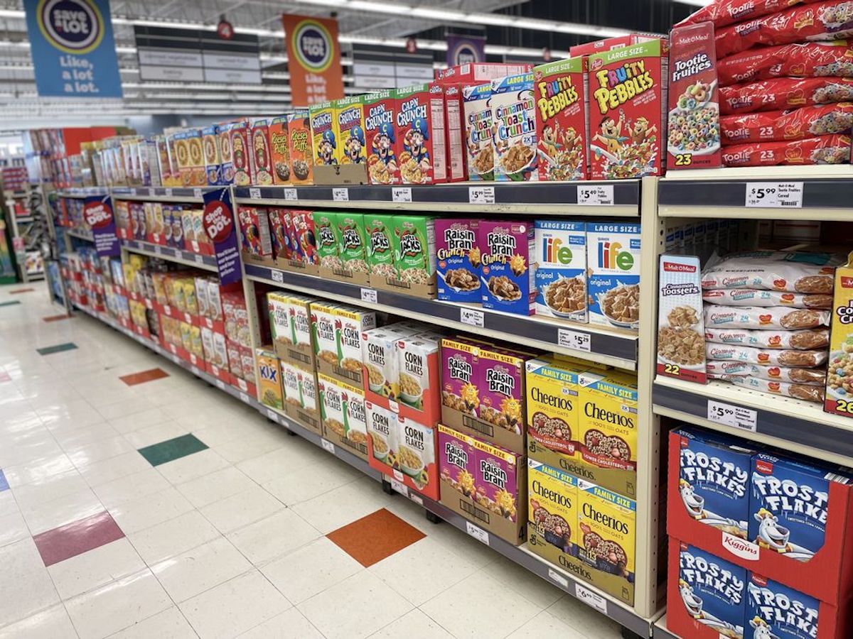 A grocery store aisle displays a variety of colorful cereal boxes, including brands like Froot Loops, Raisin Bran, Cheerios, and Frosted Flakes, neatly arranged on shelves above a checkered tile floor.
