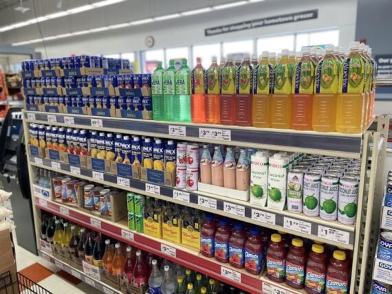 Shelves in a grocery store display various bottled drinks, including aloe vera, coconut juice, and fruit-flavored beverages, all neatly organized with visible price labels and bright packaging.
