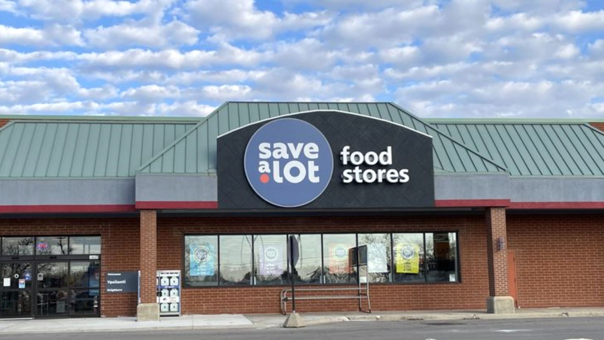 A Save A Lot food store with a green roof and a large blue and white sign above the entrance, set against a partly cloudy sky. Posters are visible in the windows below the sign.